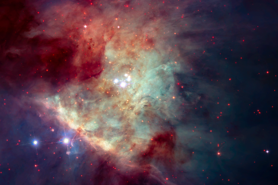 Telescope image showing a cloudy mass of various colors and brightness, a number of bright white foreground stars, and many red star-like objects in the background.