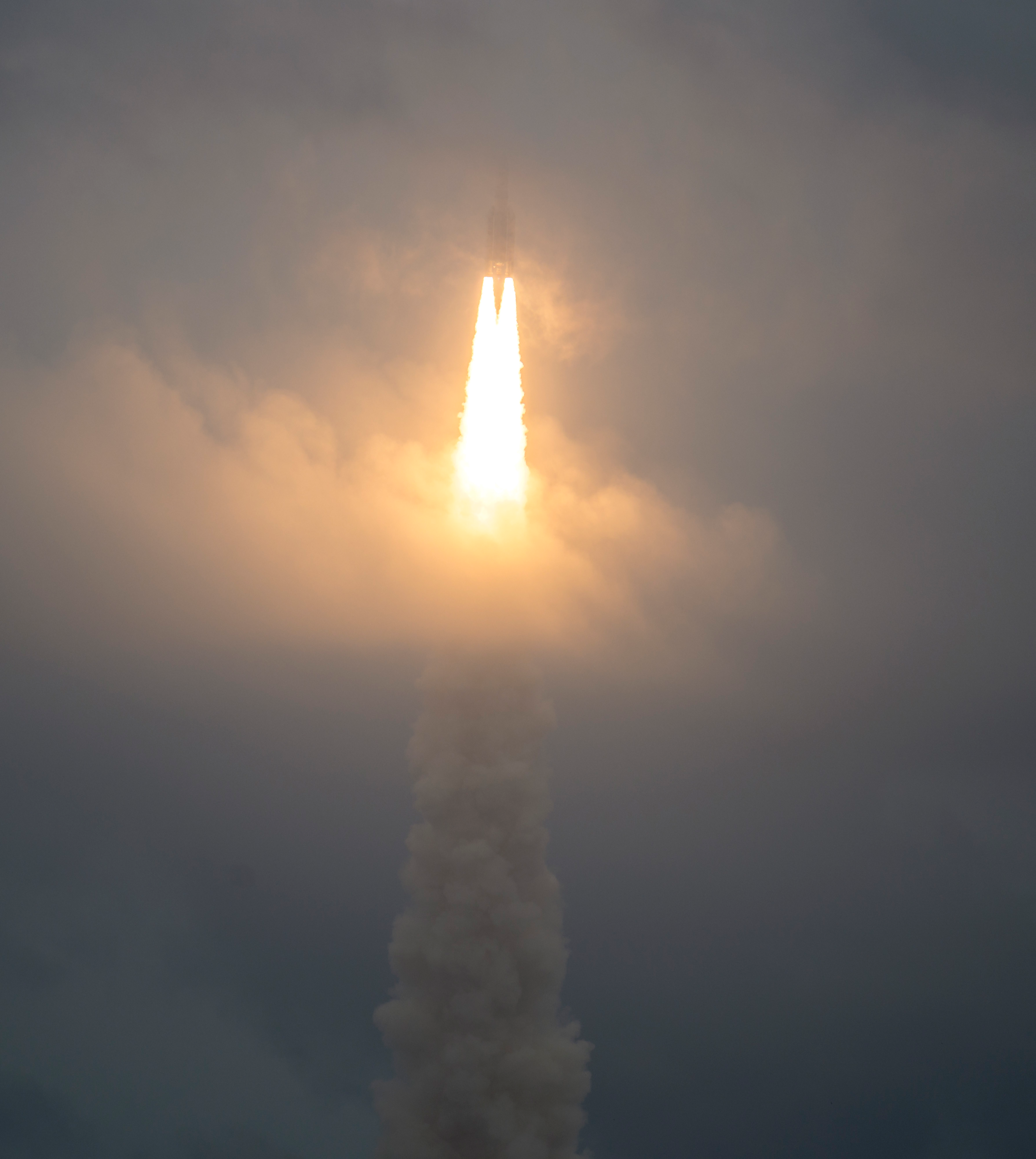 A rocket with two boosters appears very distant in the sky toward the top. The rocket pierces thick clouds, which appear orange where its boosters have recently ignited fuel. That ignition forms a bright orange triangle, which fades to shades of gray, or clouds of exhaust, at the bottom.