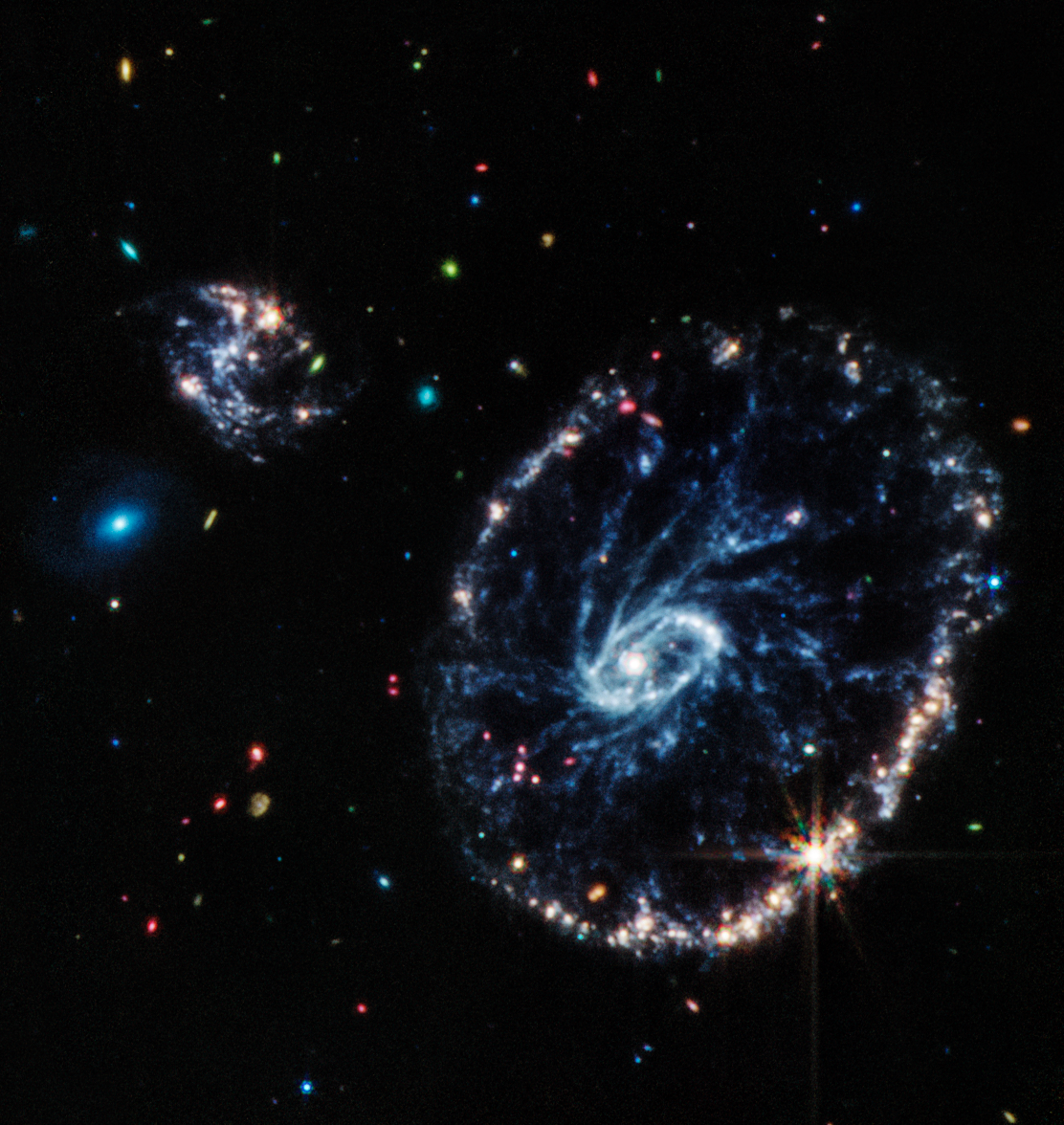 Colorful mid-infrared image of a large ring-shaped galaxy with two smaller companion galaxies, on a background of more distant galaxies of various colors, shapes, and sizes.