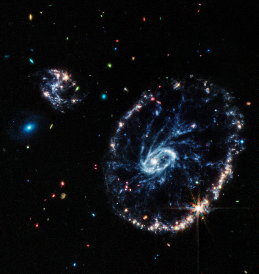Colorful mid-infrared image of a large ring-shaped galaxy with two smaller companion galaxies, on a background of more distant galaxies of various colors, shapes, and sizes.