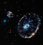 Colorful mid-infrared image of a large ring-shaped galaxy with two smaller companion galaxies, on a background of more distant galaxies of various colors, shapes, and sizes.