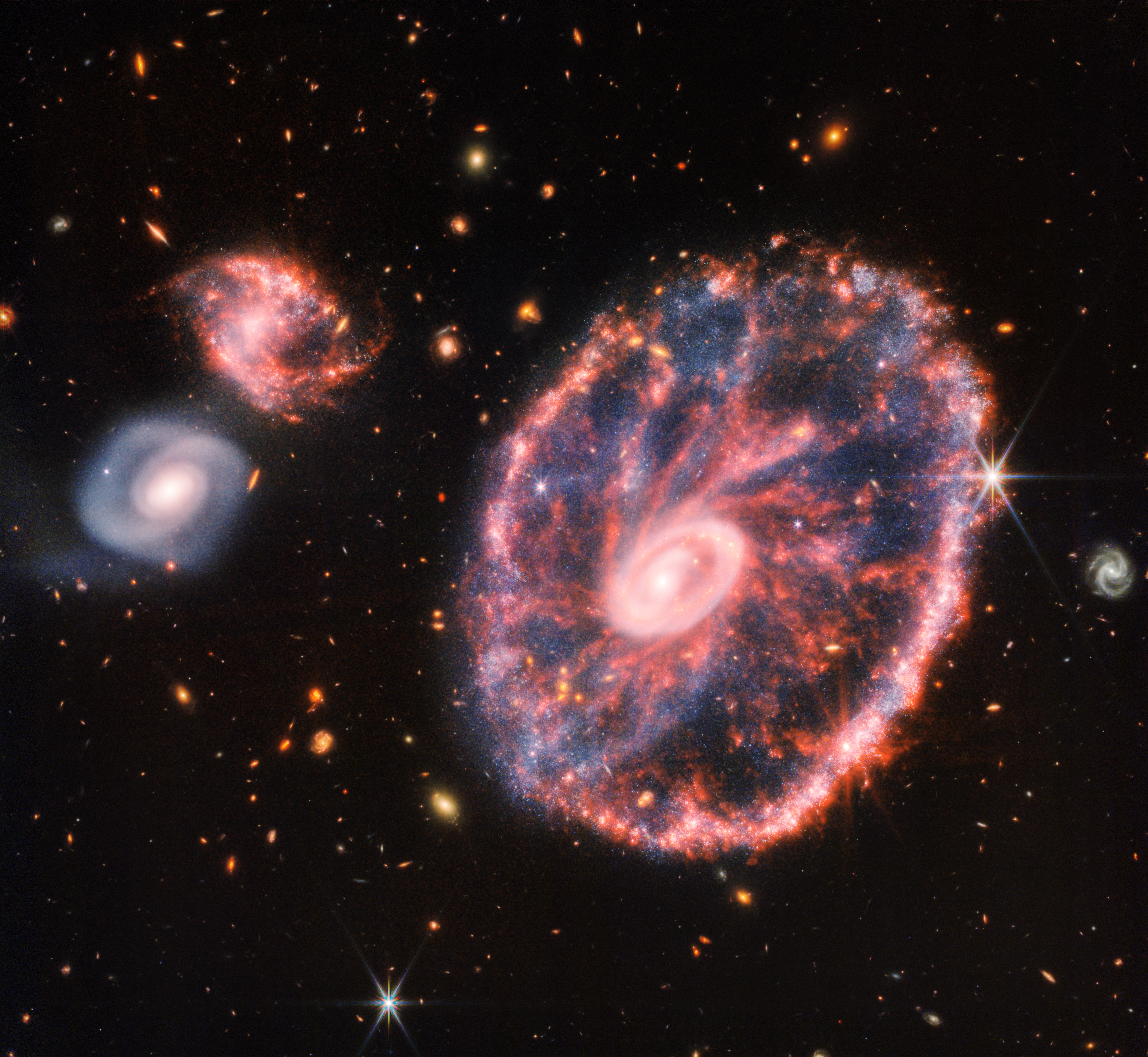Colorful near- and mid-infrared image of a large ring-shaped galaxy with two smaller spiral companion galaxies, all seen face-on, with a background of much smaller, more distant galaxies of various colors, shapes, and sizes.