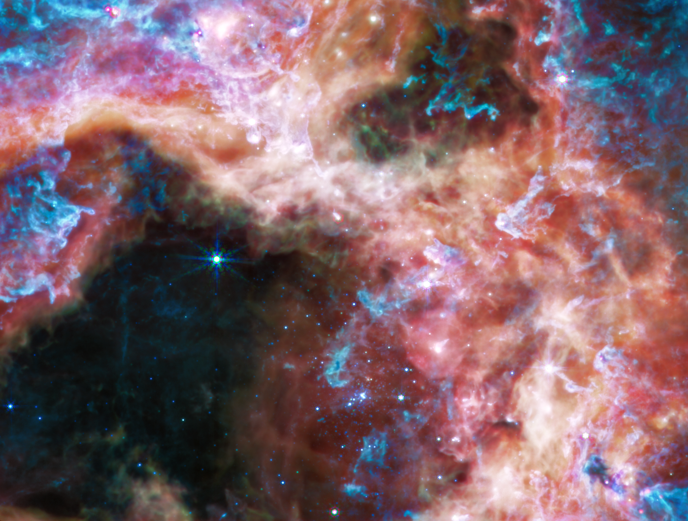 Wispy pale yellow nebula clouds highlighted with bright blue, purple and pink, surrounding a large black cavity. A few small blue stars are sprinkled at the right edge of the cavity and in the cloud. There are a few occasional bright pink spots and larger, brighter white stars. One large clump of blue dust floats amid the small blue stars. One large blue star is alone at the top of cavity, with short blue spikes and snowflake-like arms. A couple other stars in the cloud also appear like tiny snowflakes instead of points of light.