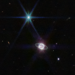 Image has a mostly dark background with one extremely bright point of light that dominates the upper left quadrant of the image and a glowing sphere towards the bottom middle of the image. The extremely bright point of light at the upper left of the image has 8 spikes pointing out from a center bright point like a compass. The glowing sphere, is mostly white, almost neon, with a few extremely bright patches of methane-ice clouds. The glowing sphere is accompanied by several narrow, faint rings and 6 tiny white dots, which are Neptune's moons. Splattered throughout the mostly black background are about 10 small, dim circles of distant galaxies.
