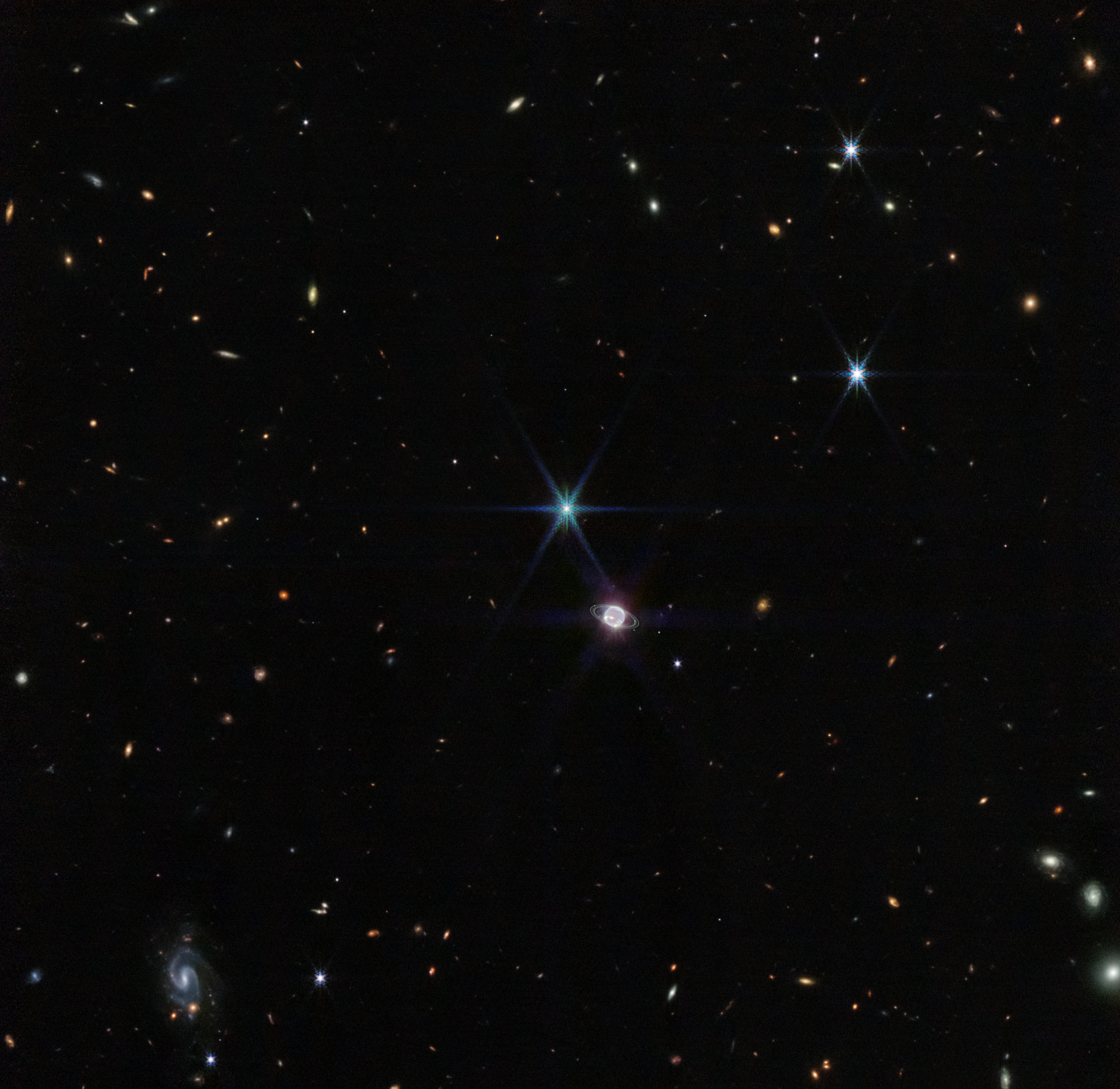 Image titled “Neptune Wide Field (NIRCam).” Hundreds of small galaxies appear around the field of view, and just a little off-center is a glowing sphere. At 11 o’clock to the glowing sphere, there is a very bright point of light with eight diffraction spikes. The colors of the galaxies that splatter across the image vary. Some are shades of orange, others are white. Most appear as fuzzy white ovals. The bright stars have diffraction spikes, forming an eight-pointed star shape. At the bottom left of the image, there is a very small barred spiral galaxy, which glows light blue.