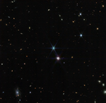 Image titled “Neptune Wide Field (NIRCam).” Hundreds of small galaxies appear around the field of view, and just a little off-center is a glowing sphere. At 11 o’clock to the glowing sphere, there is a very bright point of light with eight diffraction spikes. The colors of the galaxies that splatter across the image vary. Some are shades of orange, others are white. Most appear as fuzzy white ovals. The bright stars have diffraction spikes, forming an eight-pointed star shape. At the bottom left of the image, there is a very small barred spiral galaxy, which glows light blue.