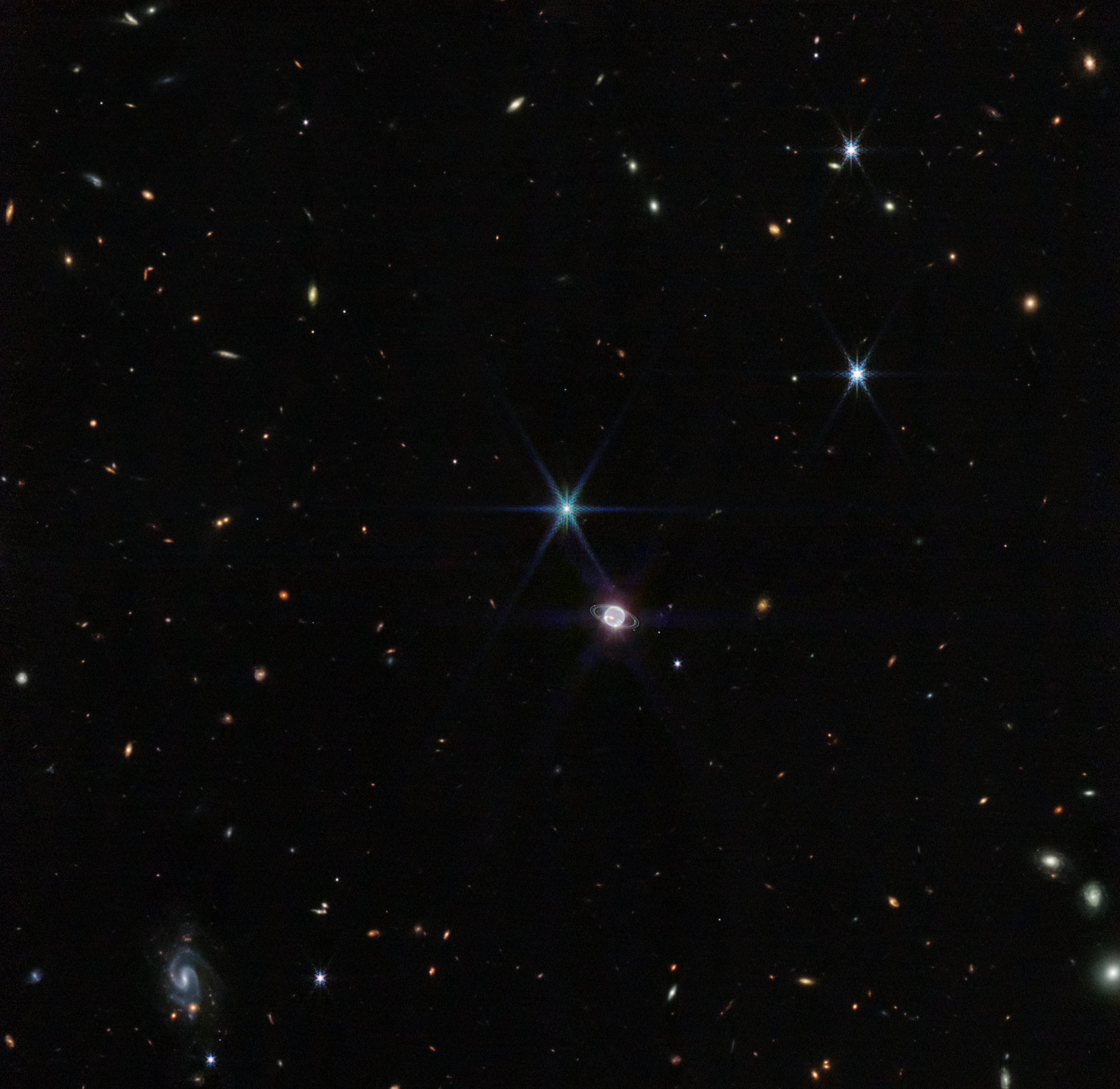 Image titled “Neptune Wide Field (NIRCam).” Hundreds of small galaxies appear around the field of view, and just a little off-center is a glowing sphere. At 11 o’clock to the glowing sphere, there is a very bright point of light with eight diffraction spikes. The colors of the galaxies that splatter across the image vary. Some are shades of orange, others are white. Most appear as fuzzy white ovals. The bright stars have diffraction spikes, forming an eight-pointed star shape. At the bottom left of the image, there is a very small barred spiral galaxy, which glows light blue.