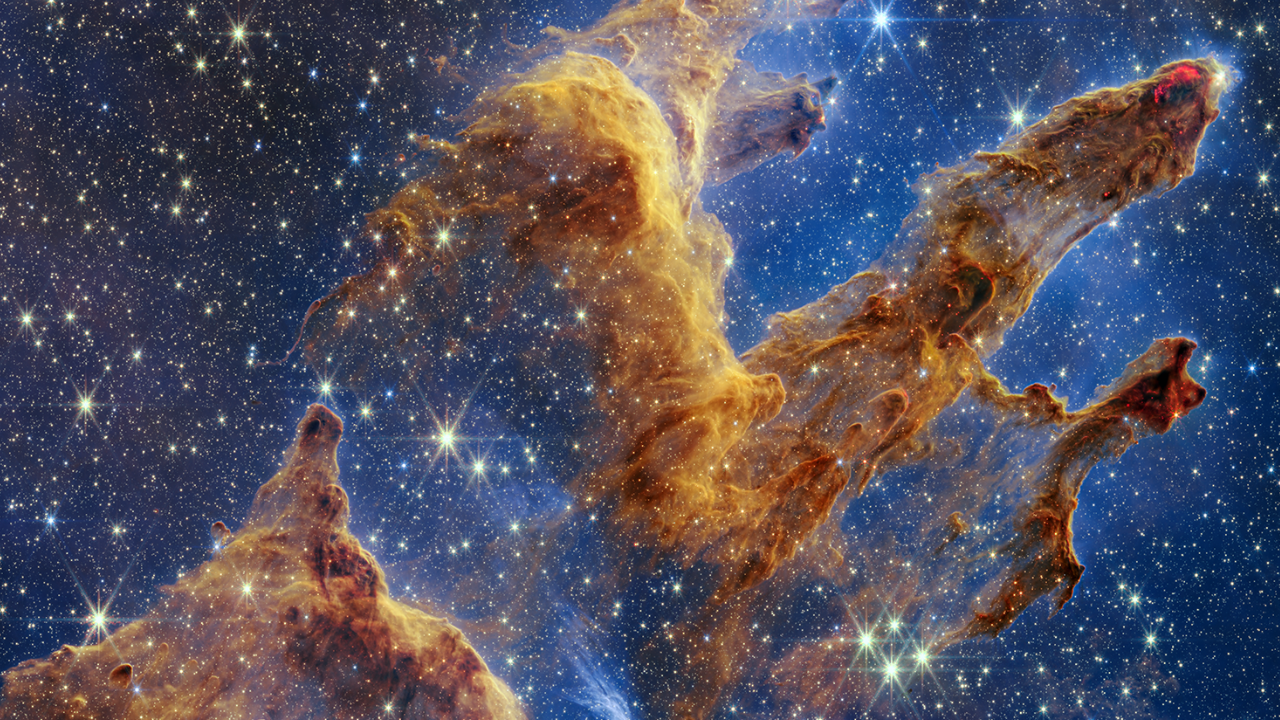 Initial video screen is split vertically. At left, the title “Webb’s New View of the Pillars of Creation” appears and at right, the near-infrared Webb image appears. The vertical image has layers of semi-opaque rusty red colored gas and dust that starts at the bottom left and goes toward the top right. There are three prominent pillars rising toward the top right. The next frame shows the center of the image close up, highlighting bright red wavy lines that look like lava along a pillar’s edge. Next, the video zooms out, showing almost the full vertical image. A quick zoom in focuses on the second pillar from the top to show the bright red orbs or stars. A screen wipe takes you to the top of the image again, to the tallest pillar, and then pans down the frame to show more of the background, which has innumerable stars. The video ends with a complete zoom out to show the full vertical image.