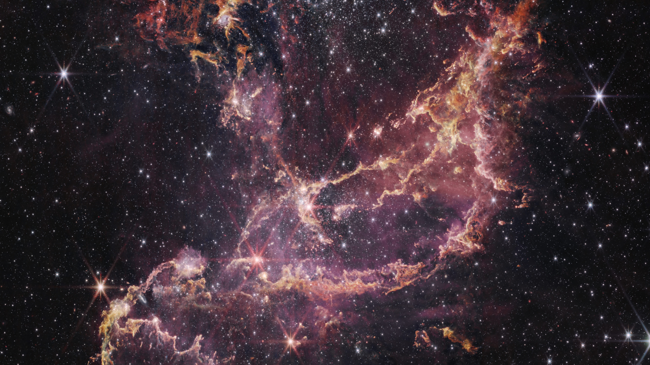 A star cluster within a nebula. The center of the image contains arcs of orange and pink gas that form a boat-like shape. One end of these arcs points to the top right of the image, while the other end points toward the bottom left. Another plume of orange and pink gas expands from the center to the top left of the image. To the right of this plume is a large cluster of white stars. There are more of these white stars and a few galaxies of different sizes spread throughout the image.