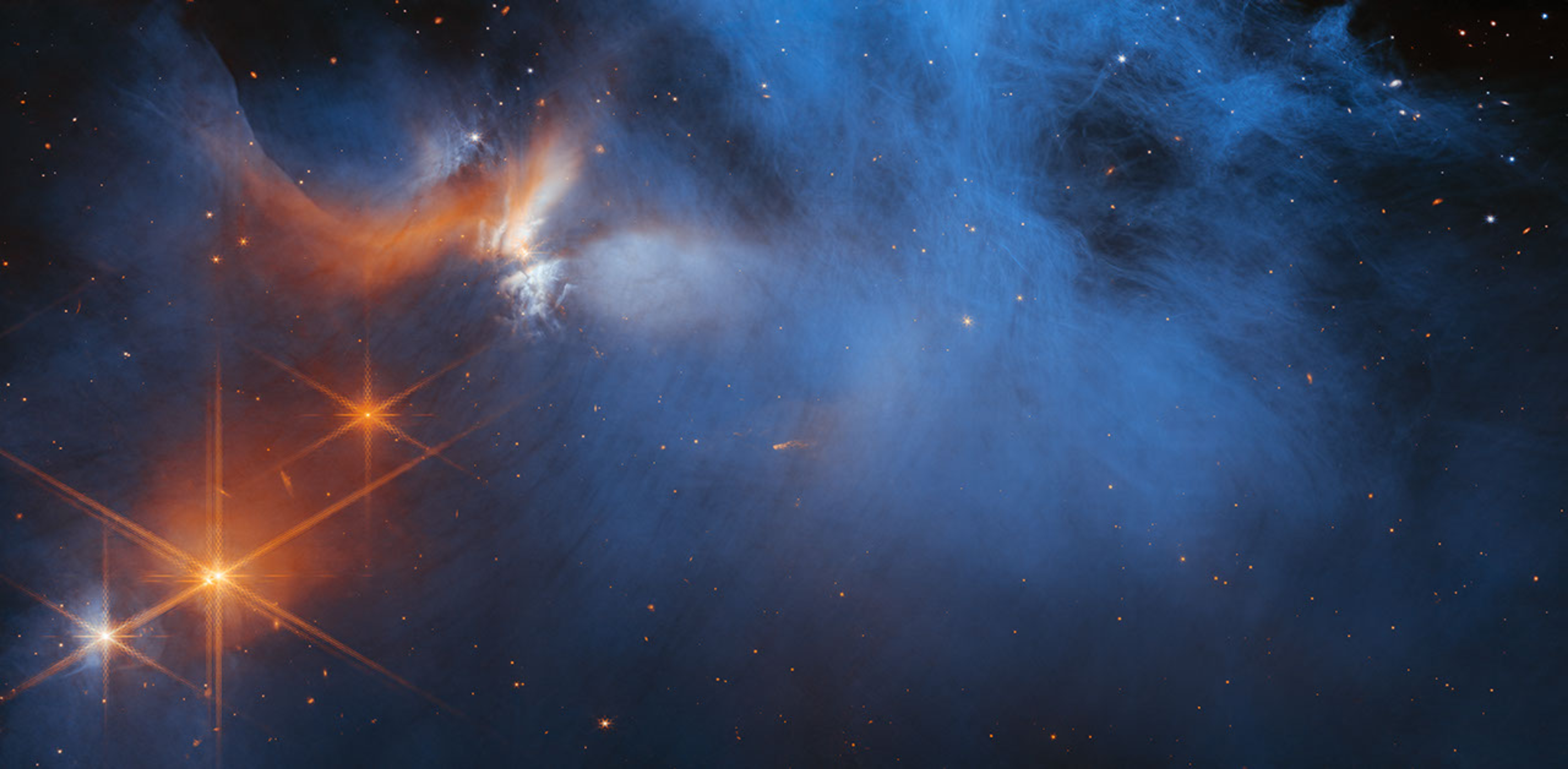 A black background is filled with pinpoints of light, which are distant galaxies, in different shades of red, orange, and blue. In the foreground and taking up most of the image are blue, smoky, translucent wisps. They begin at bottom left and make their way through the center before going out toward the top. On the left top side, some of the wisps are orange and white. Below them are four bright points of light, which are stars. Three are orange, and the one at far left appears orange-white. The stars have Webb’s signature 8-point diffraction spikes, which are also orange.