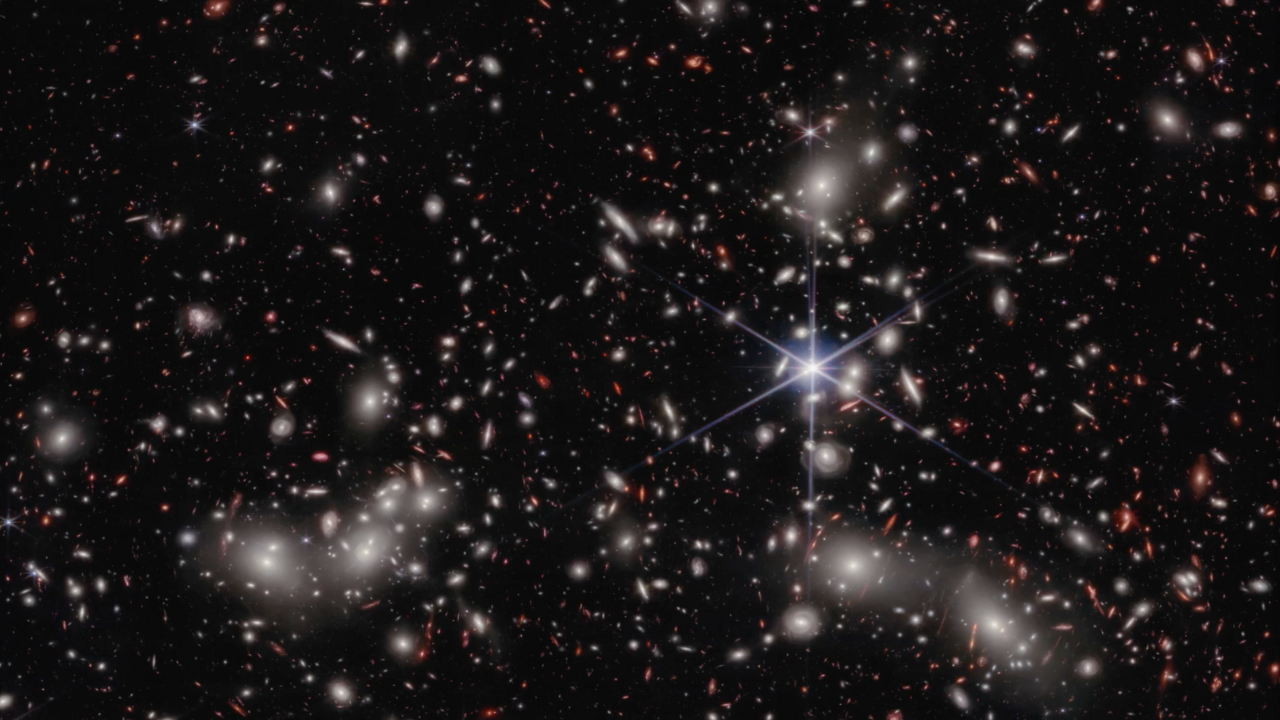 A crowded galaxy field on a black background, with one large star dominating the image just right of center. Three areas are concentrated with larger white hazy blobs on the left, lower right, and upper right above the single star. Scattered between these areas are many smaller sources of light; some also have a hazy white glow, while many other are red or orange. Even without zooming in, different galaxy shapes are detectable, like spirals, ovals, and arcs.