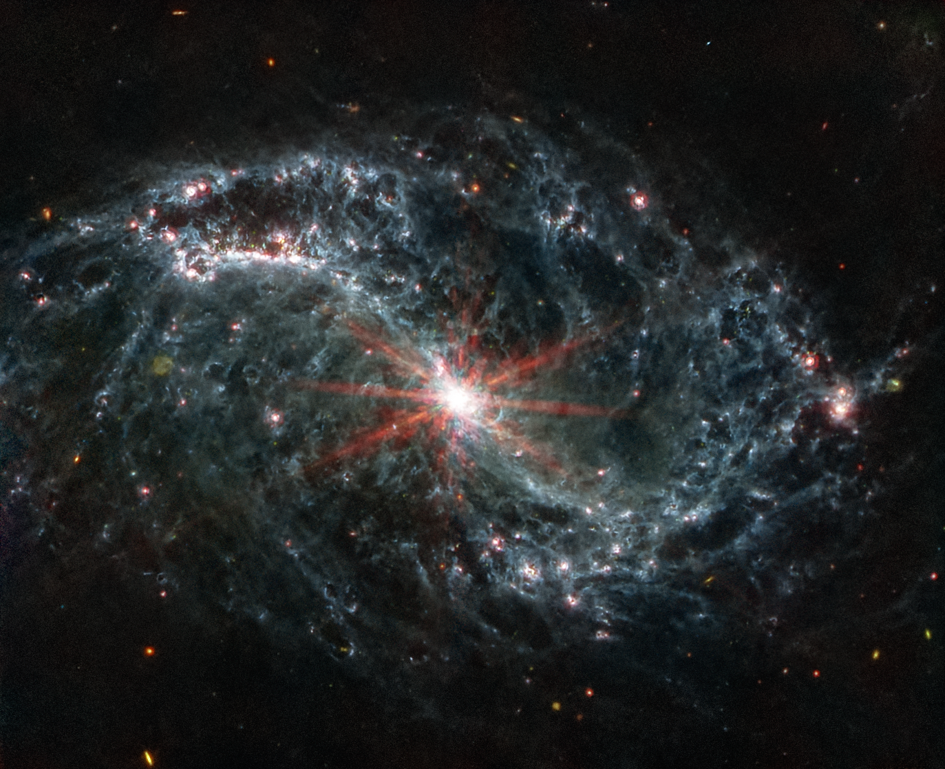 A large galaxy takes up the entirety of the image. The image is mostly black, with a bright, glowing circular core at the center. Six reddish diffraction spikes extend from the core. The spiral arms of the galaxy outstretch to the upper left and the lower right. There is black space between the core and the main spiral arms of the galaxy. Those arms are wispy and highlight filaments of dust around cavernous black bubbles. That dust in the outer rings contains diffuse dots that are navy blue, pinkish, reddish, and white. Throughout, there is also a smattering of background galaxies seen as small red and greenish dots.