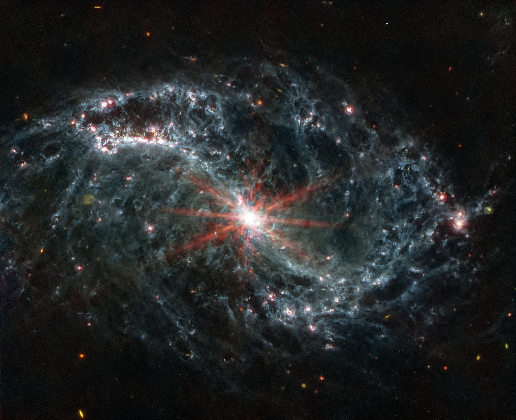 A large galaxy takes up the entirety of the image. The image is mostly black, with a bright, glowing circular core at the center. Six reddish diffraction spikes extend from the core. The spiral arms of the galaxy outstretch to the upper left and the lower right. There is black space between the core and the main spiral arms of the galaxy. Those arms are wispy and highlight filaments of dust around cavernous black bubbles. That dust in the outer rings contains diffuse dots that are navy blue, pinkish, reddish, and white. Throughout, there is also a smattering of background galaxies seen as small red and greenish dots.