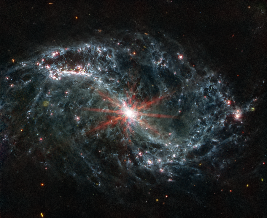 A large galaxy takes up the entirety of the image. The image is mostly black, with a bright, glowing circular core at the center. Six reddish diffraction spikes extend from the core. The spiral arms of the galaxy outstretch to the upper left and the lower right. There is black space between the core and the main spiral arms of the galaxy. Those arms are wispy and highlight filaments of dust around cavernous black bubbles. That dust in the outer rings contains diffuse dots that are navy blue, pinkish, reddish, and white. Throughout, there is also a smattering of background galaxies seen as small red and greenish dots.