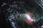 A large galaxy takes up the entirety of the image, with the lower right portion of the galaxy’s spiral arms extending off frame. The core of the galaxy is just off center to the lower right. The bright white, slightly pinkish core is not a perfect circle—it’s an elongated oval with bright, narrow, diffuse arms extending a short way out at each narrow end of the oval. The main spiral arm to the upper left of the core is wispy and highlights filaments of dust around cavernous black bubbles. Throughout, there is also a smattering of background galaxies seen as small red and greenish dots.