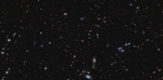 Thousands of tiny galaxies appear across the black expanse of space. The galaxy colors vary. Some of the smallest galaxies are shades of orange and pink. Most galaxies are so distant they appear as single points of light. At the center is a pink object with six diffraction spikes. This is quasar J0100+2802. It appears slightly smaller than the foreground stars, which appear blue.