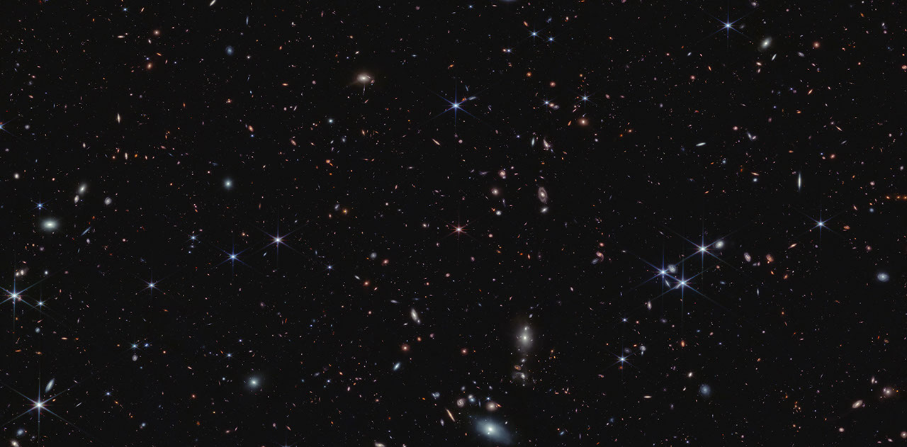 Thousands of tiny galaxies appear across the black expanse of space. The galaxy colors vary. Some of the smallest galaxies are shades of orange and pink. Most galaxies are so distant they appear as single points of light. At the center is a pink object with six diffraction spikes. This is quasar J0100+2802. It appears slightly smaller than the foreground stars, which appear blue.