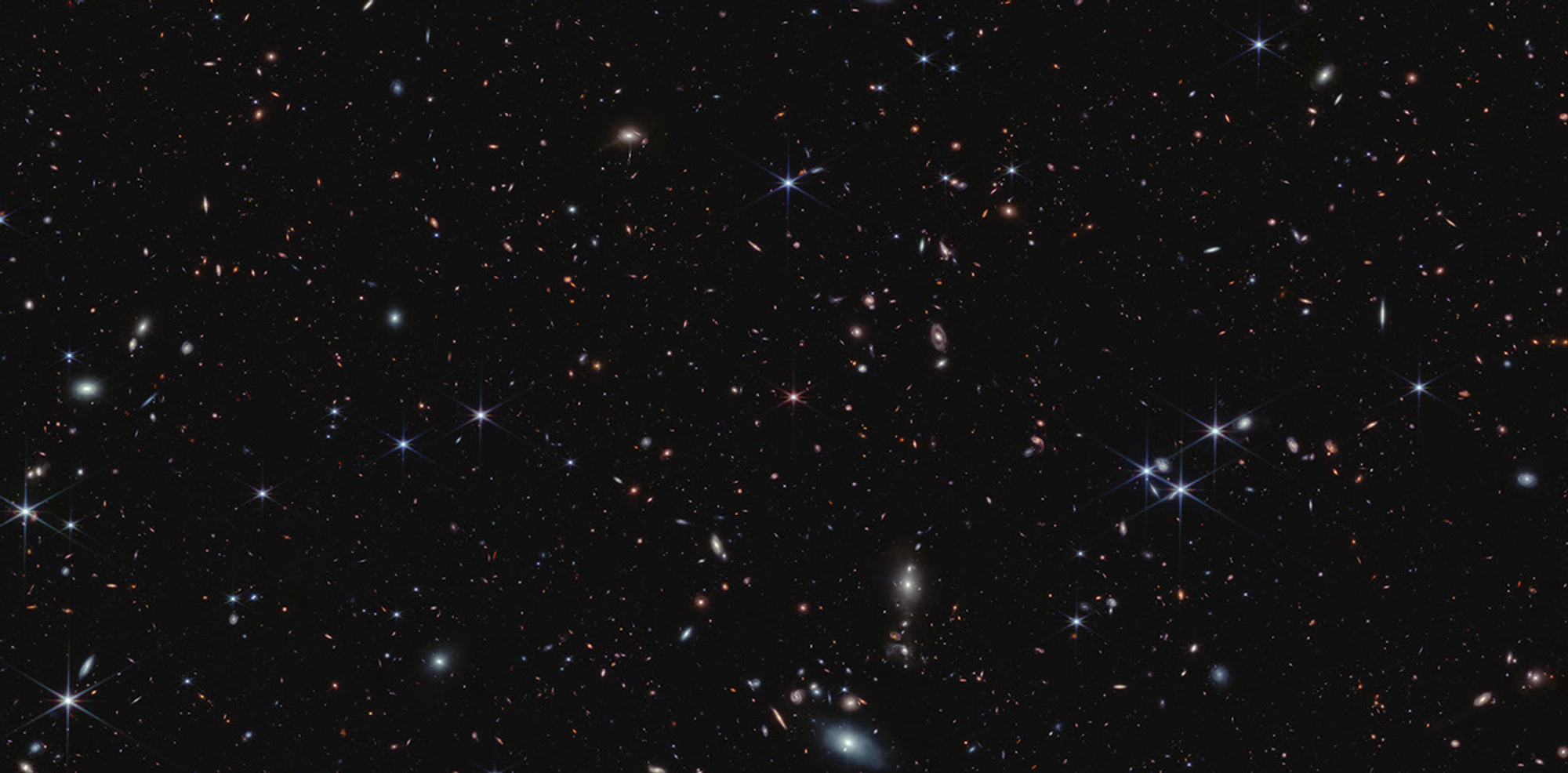 Thousands of tiny galaxies appear across the black expanse of space. The galaxy colors vary. Some of the smallest galaxies are shades of orange and pink. Most galaxies are so distant they appear as single points of light. At the center is a pink object with six diffraction spikes. This is quasar J0100+2802. It appears slightly smaller than the foreground stars, which appear blue.