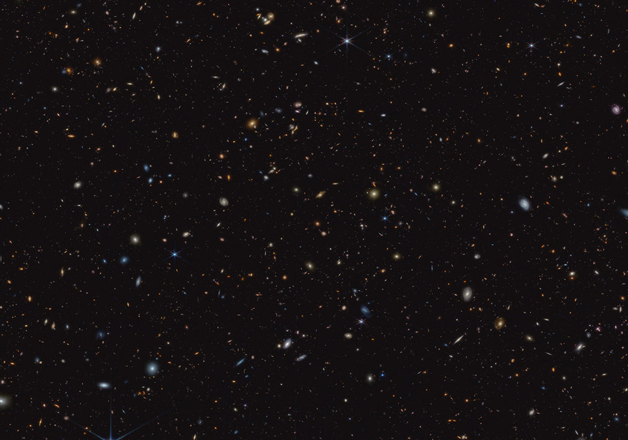 Thousands of small galaxies are scattered on a black background. Some are noticeably spirals, either face-on or edge-on, while others are blobby ellipticals. Many are too small to discern any structure. A few spirals are bluish, but most of the galaxies appear yellow or red. A handful of stars display eight-point diffraction spikes.