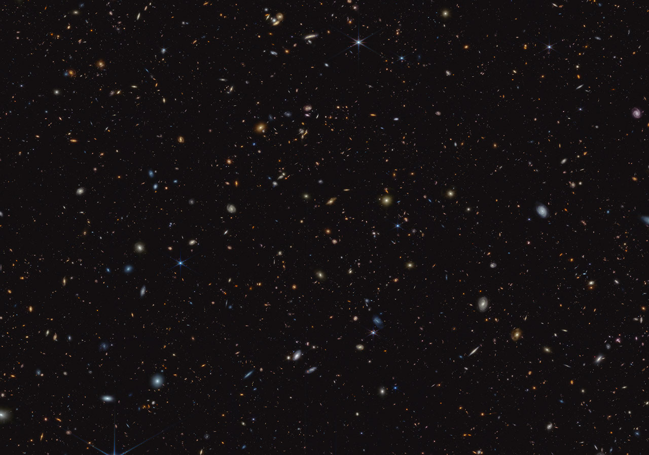 Thousands of small galaxies are scattered on a black background. Some are noticeably spirals, either face-on or edge-on, while others are blobby ellipticals. Many are too small to discern any structure. A few spirals are bluish, but most of the galaxies appear yellow or red. A handful of stars display eight-point diffraction spikes.