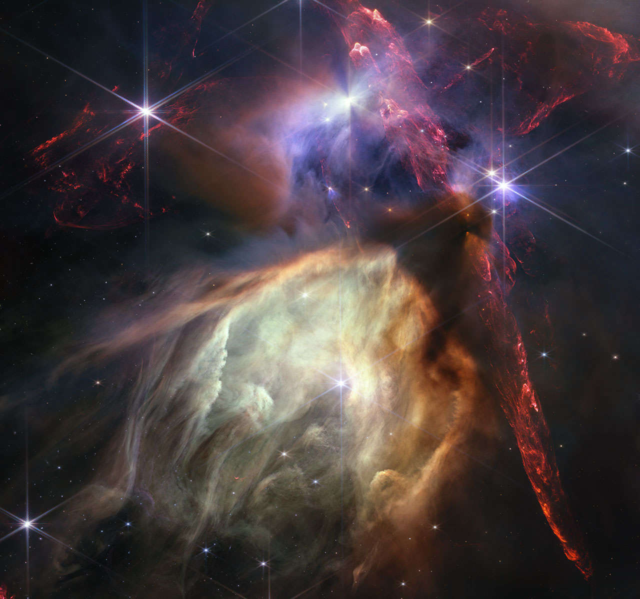 Red dual opposing jets coming from young stars fill the darker top half of the image, while a glowing pale-yellow, cave-like structure is bottom center, tilted toward two o’clock, with a bright star at its center. The dust of the cave structure becomes wispy toward eight o’clock. Above the arched top of the dust cave three groupings of stars with diffraction spikes are arranged. A dark cloud sits at the top of the arch of the glowing dust cave, with one streamer curling down the right-hand side. The dark shadow of the cloud appears pinched in the center, with light emerging in a triangle shape above and below the pinch, revealing the presence of a star inside the dark cloud. The image’s largest jets of red material emanate from within this dark cloud, thick and displaying structure like the rough face of a cliff, glowing brighter at the edges. At the top center of the image, a star displays another, larger pinched dark shadow, this time vertically. To the left of this star is a more wispy, indistinct region.
