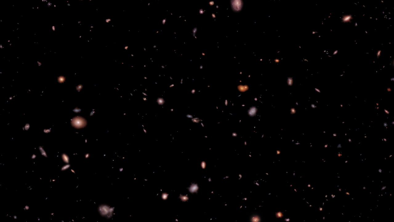 An image of thousands of galaxies of different shapes and sizes in different shades of red, yellow, blue, and white. Most of these galaxies are fuzzy ovals with bright centers, while others appear like bright streaks atop the black background of space.