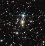 A black background is scattered with hundreds of small galaxies of different shapes, ranging in color from white to yellow to red. Some galaxies, mostly the redder galaxies, are distorted, appearing to be stretched out or mirror imaged. Just a little bit above the center, there is a bright source of light, a star, with 8 bright diffraction spikes extending out from it. Below the star are several noticeably fuzzy white galaxies that resemble cotton balls – these are part of a galaxy cluster. To the lower right of the star is a particularly long, red, thin line stretching from one o’clock to 7 o’clock. This is the Sunrise Arc.