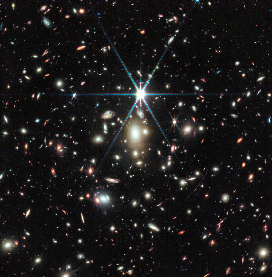 A black background is scattered with hundreds of small galaxies of different shapes, ranging in color from white to yellow to red. Some galaxies, mostly the redder galaxies, are distorted, appearing to be stretched out or mirror imaged. Just a little bit above the center, there is a bright source of light, a star, with 8 bright diffraction spikes extending out from it. Below the star are several noticeably fuzzy white galaxies that resemble cotton balls – these are part of a galaxy cluster. To the lower right of the star is a particularly long, red, thin line stretching from one o’clock to 7 o’clock. This is the Sunrise Arc.