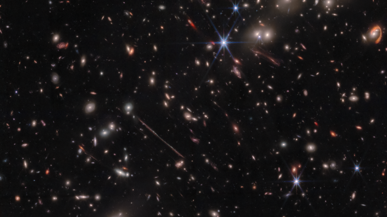 A black background is scattered with hundreds of small galaxies of different shapes, ranging in color from white to yellow to red. Some galaxies are distorted, appearing to be stretched out or mirror imaged. Near the center, a particularly long and thin line stretches from ten o’clock to four o’clock. At upper right, a red swoosh extends about three-quarters of the way around a pair of galaxies. A handful of foreground stars display eight diffraction spikes.