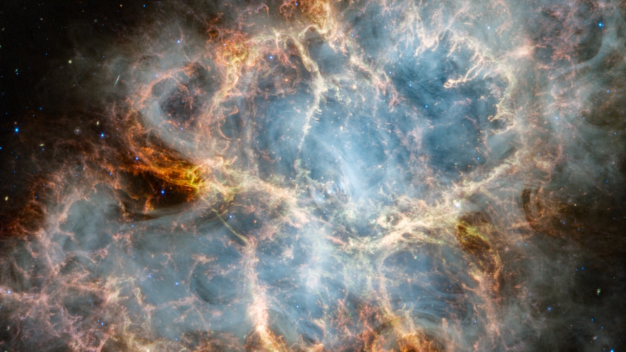 The Crab Nebula. An oval nebula with complex structure against a black background. On the nebula’s exterior, particularly at the top left and bottom left, lie curtains of glowing red and orange fluffy material. Its interior shell shows large-scale loops of mottled filaments of yellow-white and green, studded with clumps and knots. Translucent thin ribbons of smoky white lie within the remnant’s interior, brightest toward its center. The white material follows different directions throughout, including sometimes sharply curving away from certain regions within the remnant. A faint, wispy ring of white material encircles the very center of the nebula. Around and within the supernova remnant are many points of blue, red, and yellow light.