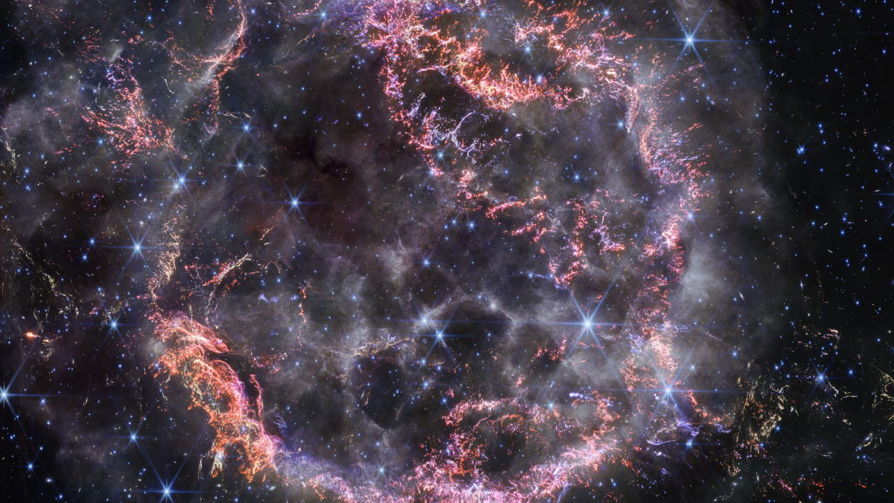 Cassiopeia A, a circular-shaped cloud of gas and dust with complex structure. The inner shell is made of bright pink and orange filaments studded with clumps and knots that look like tiny pieces of shattered glass. Around the exterior of the inner shell, particularly at the upper right, there are curtains of wispy gas that look like campfire smoke. The white smoke-like material also appears to fill the cavity of the inner shell, featuring structures shaped like large bubbles. Around and within the nebula, there are various stars seen as points of blue and white light.