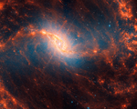 Webb’s image of galaxy NGC 1365 shows a central region that looks like an angled, smashed oval that is a mix of bright orange shades that emanate a blue glow. The galaxy’s bar extends horizontally from the oval. It is crossed by messy dust filaments that curve slightly, forming a backward S shape. Fainter filaments appear below and above the core.