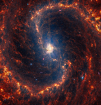 Webb’s image of NGC 4535 shows a densely populated face-on barred spiral galaxy anchored by its central region, which has a light orange haze. The galaxy’s core is centered and connected by an almost vertical bar structure to the galaxy’s two prominent spiral arms, which appear to rotate clockwise, forming an elongated S shape in shades of orange.