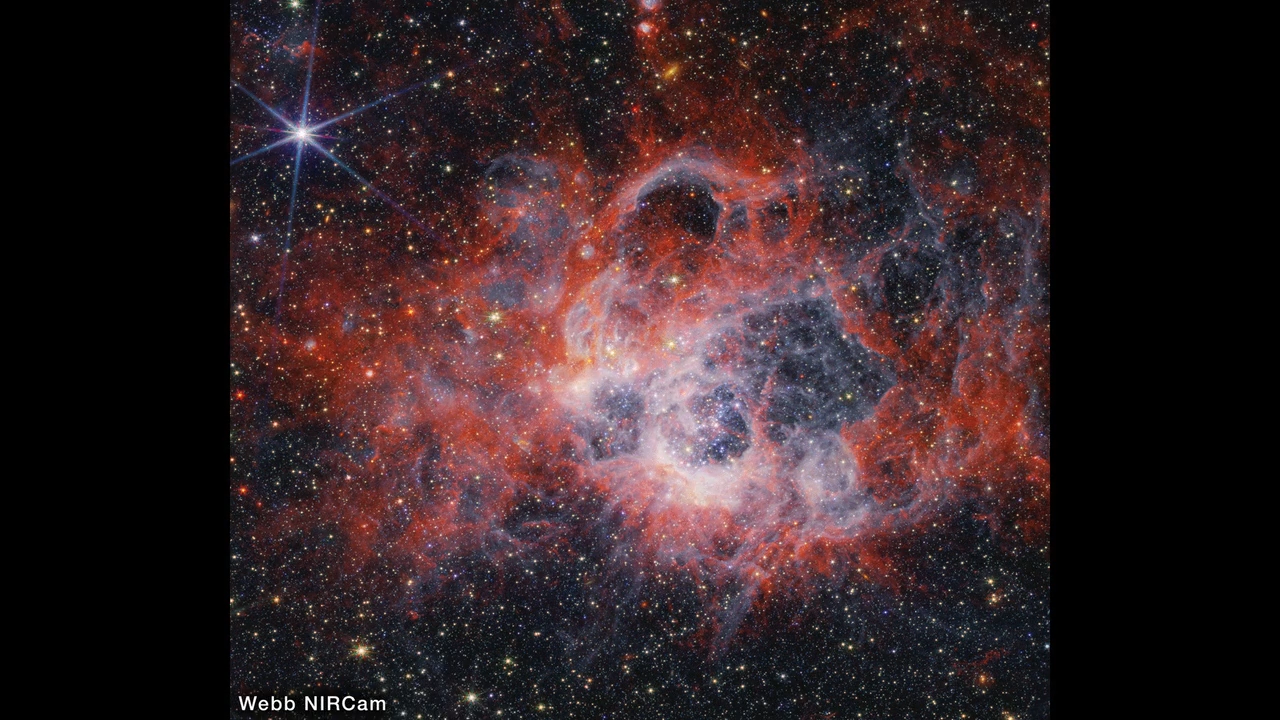 At the center of the image is a nebula on the black background of space. The nebula is comprised of clumpy, red, filamentary clouds. At the center-right of the red clouds is a large cavernous bubble, and at the center of the bubble there is a opaque blueish glow with speckles of stars. At the edges of the bubble, the dust is white. There are several other smaller cavernous bubbles at the top of the nebula. There are thousands of stars that fill the surrounding area outside of the nebula, most of them are yellow or white – at 11 o’clock and 6 o’clock there are extremely bright stars with 8 diffraction spikes. There are also some smaller, red stars and a few disc-shaped galaxies scattered about the image.