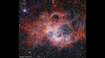 At the center of the image is a nebula on the black background of space. The nebula is comprised of clumpy, red, filamentary clouds. At the center-right of the red clouds is a large cavernous bubble, and at the center of the bubble there is a opaque blueish glow with speckles of stars. At the edges of the bubble, the dust is white. There are several other smaller cavernous bubbles at the top of the nebula. There are thousands of stars that fill the surrounding area outside of the nebula, most of them are yellow or white – at 11 o’clock and 6 o’clock there are extremely bright stars with 8 diffraction spikes. There are also some smaller, red stars and a few disc-shaped galaxies scattered about the image.