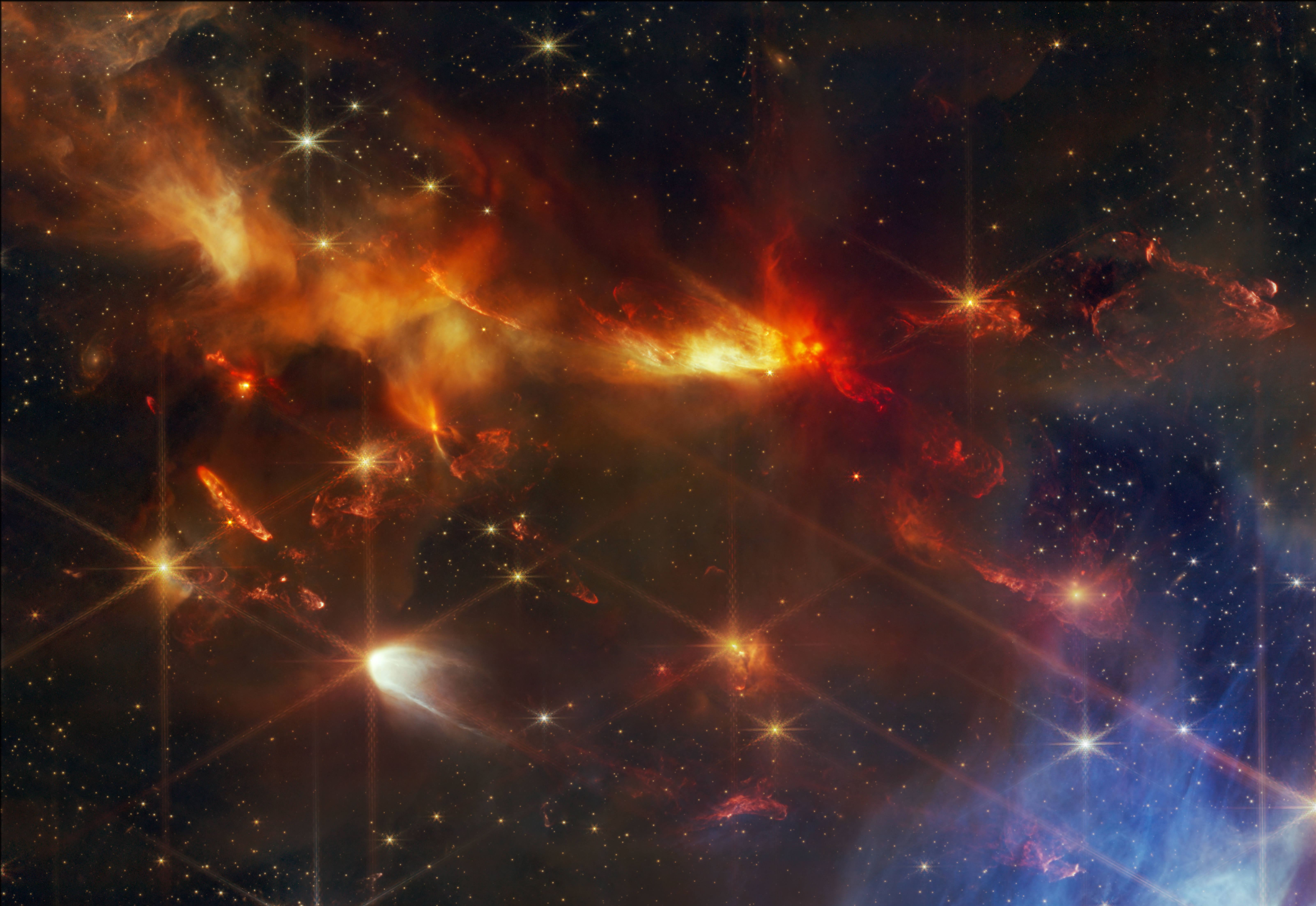 A portion of the young star-forming region Serpens Nebula. It’s filled with wispy orange and red layers of gas and dust, and within that orange dust, there are several small red plumes of gas that extend from the top left to the bottom right, at the same angle. There are wispy blue filaments of glass in the bottom right corner of the image. Small points of light are sprinkled across the field, brightest sources in the field have extensive eight-pointed diffraction spikes that are characteristic of the Webb Telescope.