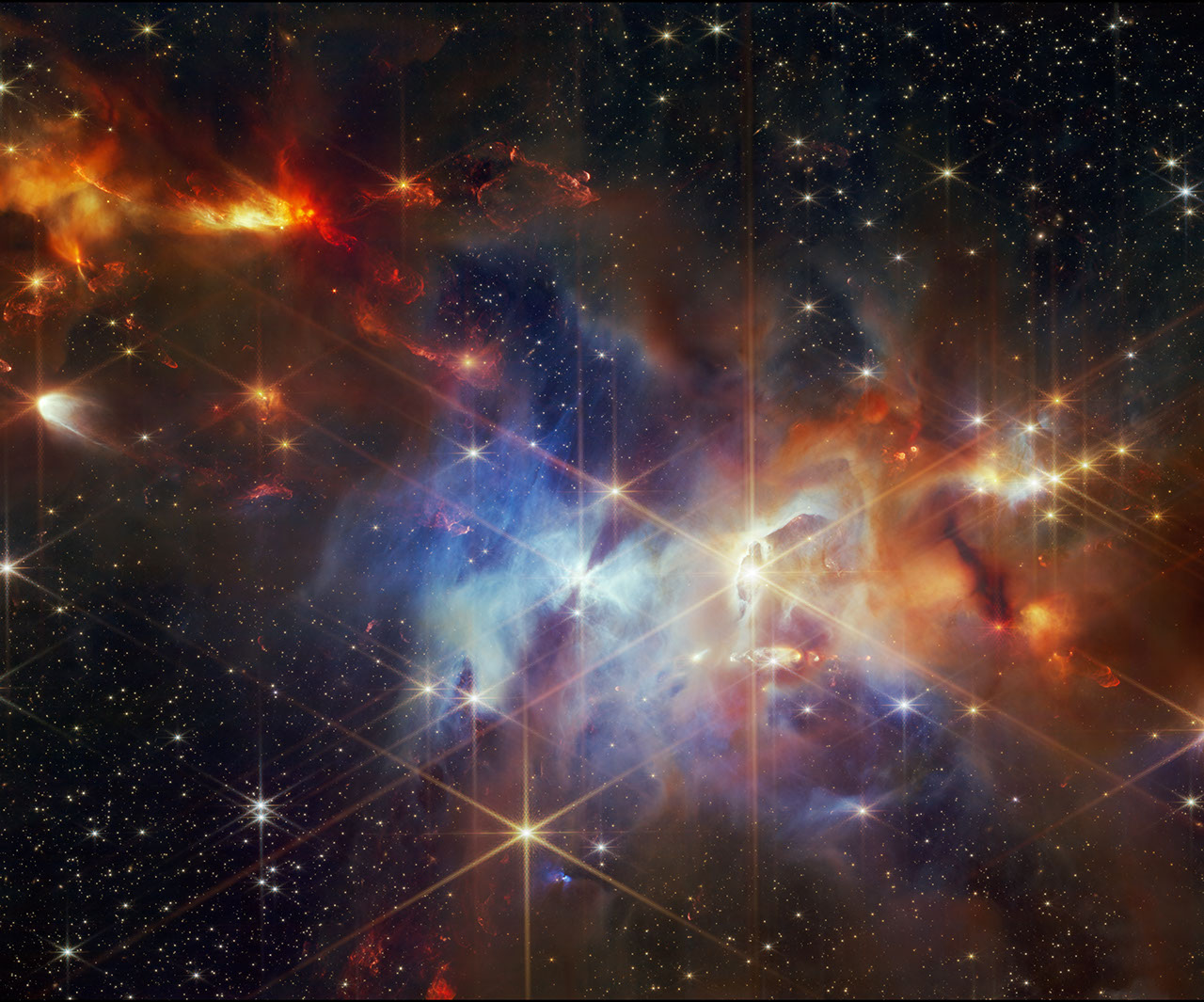 A young star-forming region is filled with wispy orange, red, and blue layers of gas and dust. The upper left corner of the image is filled with mostly orange dust, and within that orange dust, there are several small red plumes of gas that extend from the top left to the bottom right, at the same angle. The center of the image is filled with mostly blue gas. At the center, there is one particularly bright star, that has an hourglass shadow above and below it. To the right of that is what looks a vertical eye-shaped crevice with a bright star at the center. The gas to the right of the crevice is a darker orange. Small points of light are sprinkled across the field, brightest sources in the field have extensive eight-pointed diffraction spikes that are characteristic of the Webb Telescope.