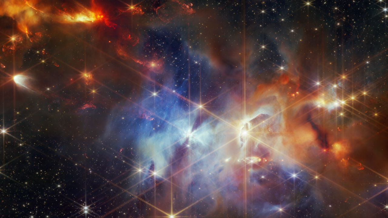 A young star-forming region is filled with wispy orange, red, and blue layers of gas and dust. The upper left corner of the image is filled with mostly orange dust, and within that orange dust, there are several small red plumes of gas that extend from the top left to the bottom right, at the same angle. The center of the image is filled with mostly blue gas. At the center, there is one particularly bright star, that has an hourglass shadow above and below it. To the right of that is what looks a vertical eye-shaped crevice with a bright star at the center. The gas to the right of the crevice is a darker orange. Small points of light are sprinkled across the field, brightest sources in the field have extensive eight-pointed diffraction spikes that are characteristic of the Webb Telescope.