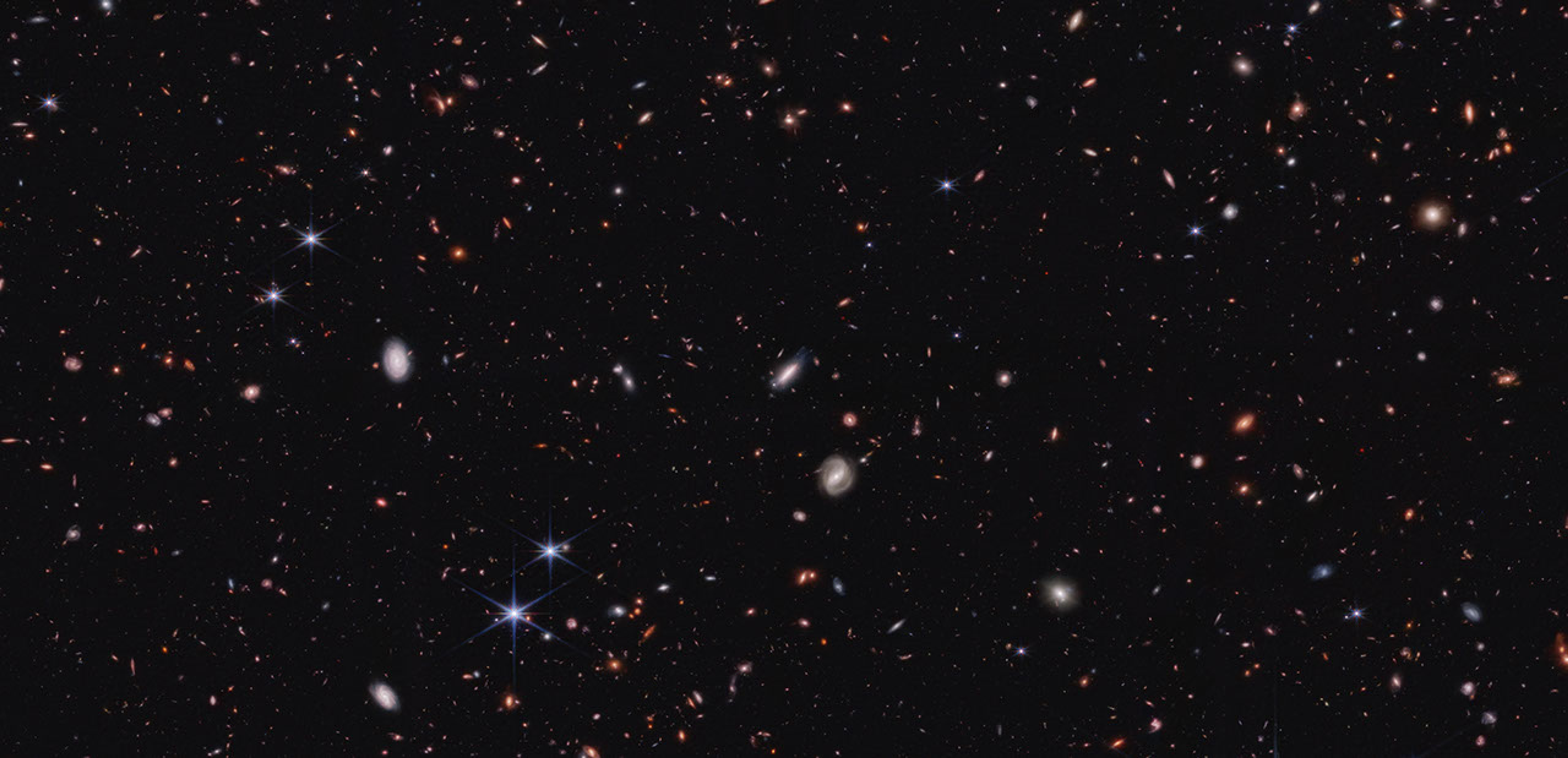 Hundreds of small galaxies against the black background of space. Several white spiral galaxies are near image center. Most of the galaxies are various shades of orange and red, and many are too tiny to discern a shape. A handful of foreground stars show Webb's six diffraction spikes.