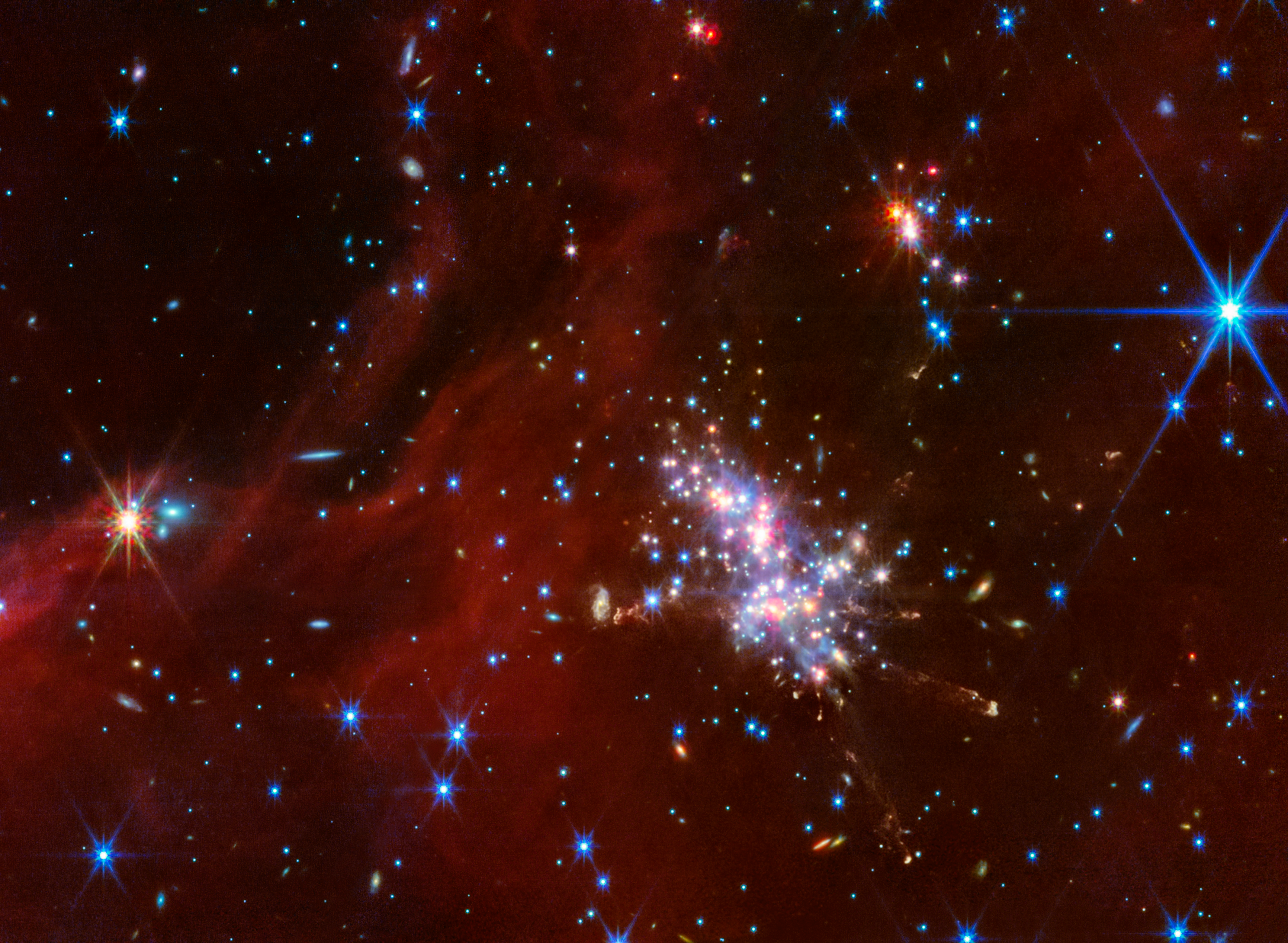 At center right is a compact star cluster composed of luminous red, blue, and white points of light. Faint jets with clumpy, diffuse material extend in various directions from the bright cluster. Above and to the right is a smaller cluster of stars. Translucent red wisps of material stretch across the scene. Background galaxies are scattered across this swath of space, appearing as small blue-white and orange-white dots or fuzzy, thin disks.