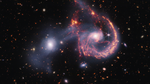 A pair of interacting galaxies. The larger of the two galaxies is slightly right of center, and composed of a hazy, bright, white center and a ring of gaseous filaments, which are different shades of red and orange. Toward the bottom left and bottom right of the ring are filaments of gas spiraling inward toward the core. At the top left of the ring is a noticeable gap, bordered by two large, orange pockets of dust and gas. The smaller galaxy to its left is made of hazy white gas and dust, which becomes more diffuse farther away from its center. To this galaxy’s bottom left, there is a smaller, more diffuse gas cloud that wafts outward toward the edges. Many red, orange, and white galaxies are spread throughout, with some hazier in composition and others having more defined spiral patterns.