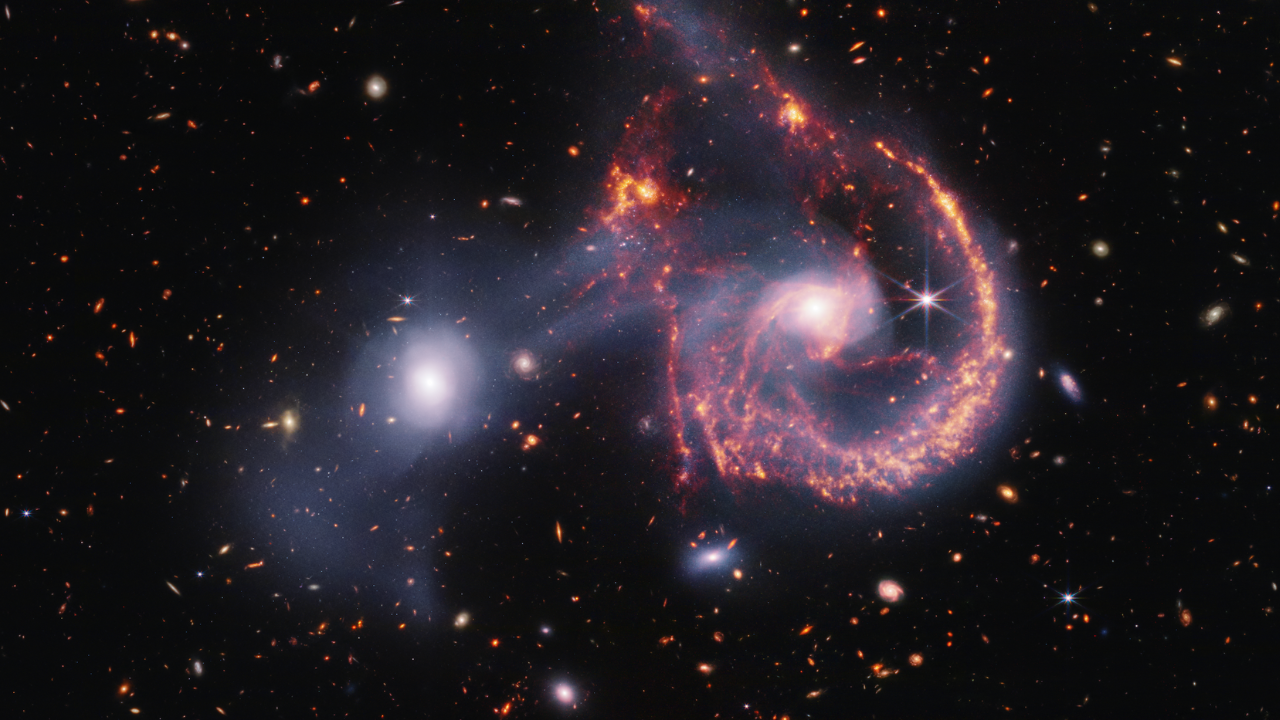 A pair of interacting galaxies. The larger of the two galaxies is slightly right of center, and composed of a hazy, bright, white center and a ring of gaseous filaments, which are different shades of red and orange. Toward the bottom left and bottom right of the ring are filaments of gas spiraling inward toward the core. At the top left of the ring is a noticeable gap, bordered by two large, orange pockets of dust and gas. The smaller galaxy to its left is made of hazy white gas and dust, which becomes more diffuse farther away from its center. To this galaxy’s bottom left, there is a smaller, more diffuse gas cloud that wafts outward toward the edges. Many red, orange, and white galaxies are spread throughout, with some hazier in composition and others having more defined spiral patterns.