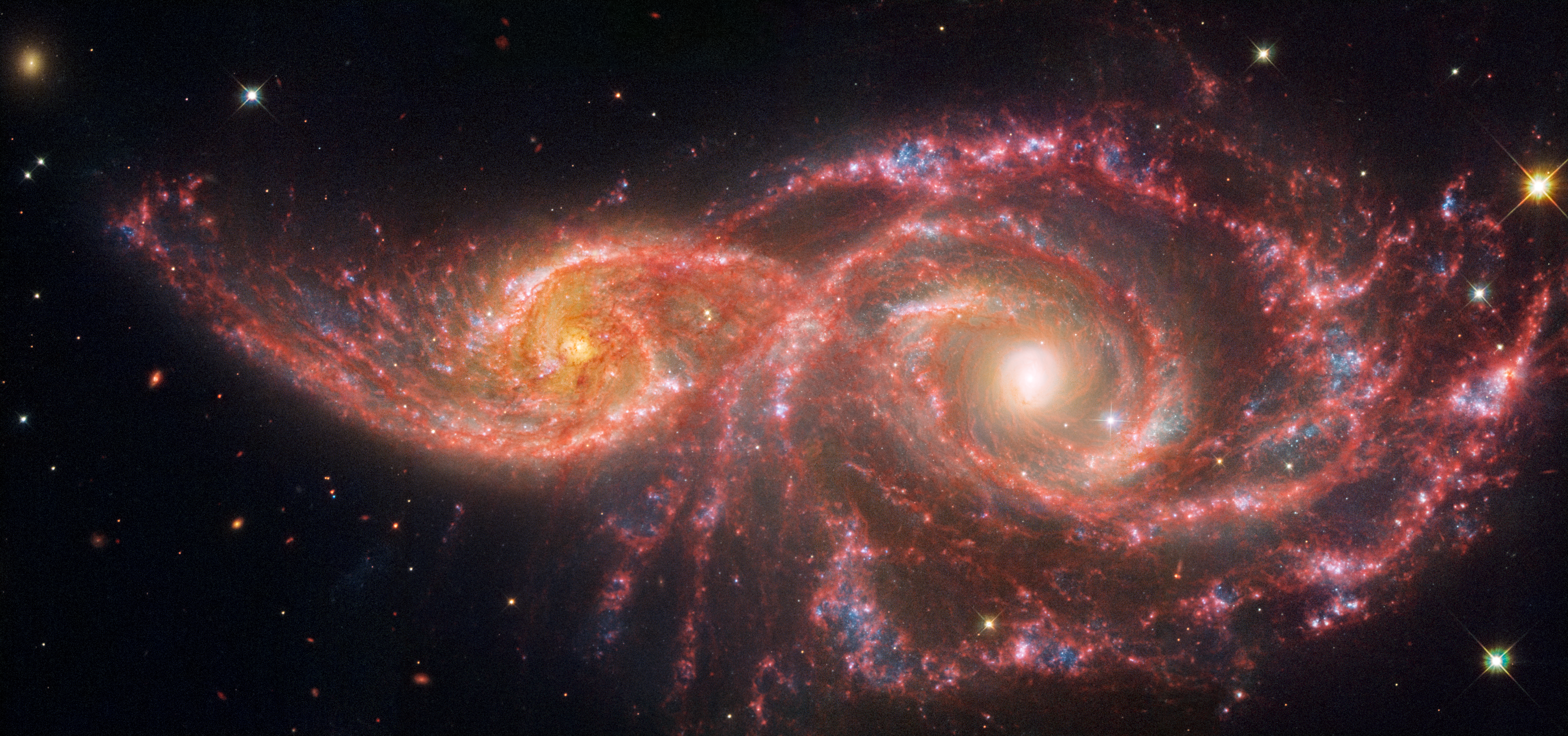 Two spiral galaxies take up almost the entire view and appear to be overlapping. The galaxy at left, IC 2163, is smaller and more compact than the galaxy at right, NGC 2207. The black background of space is dotted with foreground stars and extremely distant galaxies.