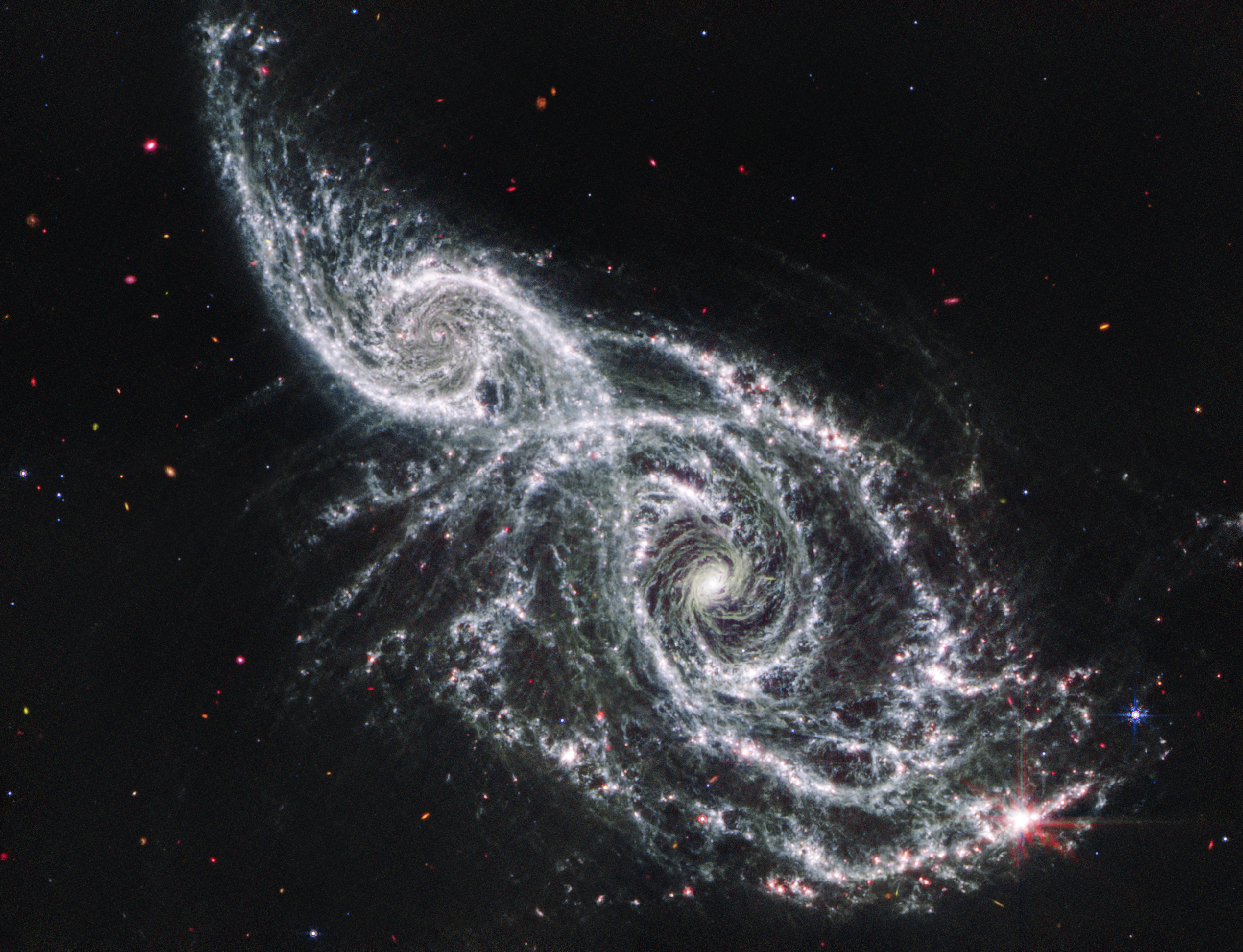 Two spiral galaxies take up almost the entire view and appear to be overlapping. They are angled from top left to bottom right. The galaxy at left, IC 2163, is smaller and more compact than the galaxy at right, NGC 2207. The background of space is black, dotted with tiny foreground stars and extremely distant galaxies.