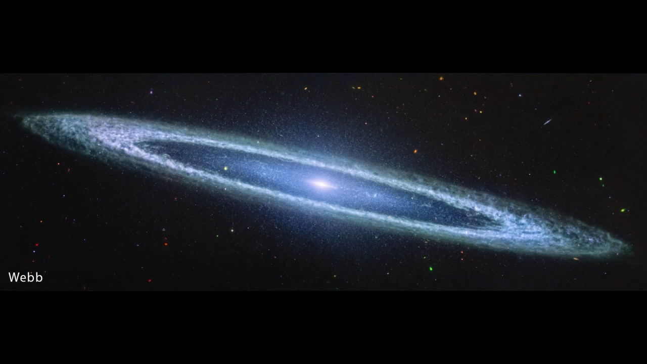 Image of a galaxy on the black background of space. The galaxy is a very oblong, blue disk that extends from left to right at an angle (from about 10 o’clock to 5 o’clock). The galaxy has a small bright core at the center. There is an inner disk that is clearer, with speckles of stars scattered throughout. The outer disk of the galaxy is whiteish-blue, and clumpy, like clouds in the sky. There are different colored dots, distant galaxies, speckled among the black background of space surrounding the galaxy.