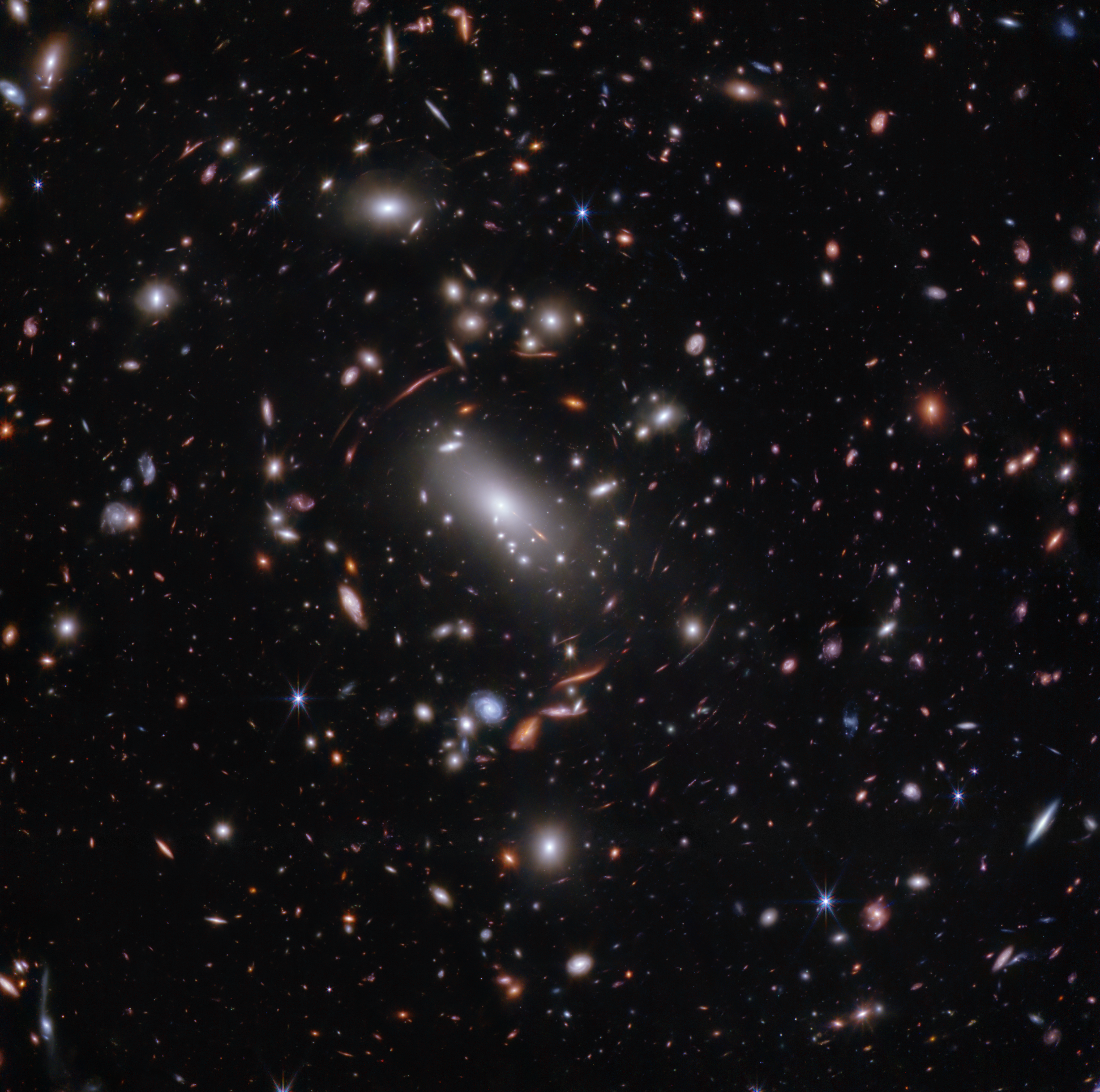 Thousands of overlapping objects at various distances are spread across this field, including galaxies in a massive galaxy cluster, and distorted background galaxies behind the galaxy cluster. The background of space is black.
