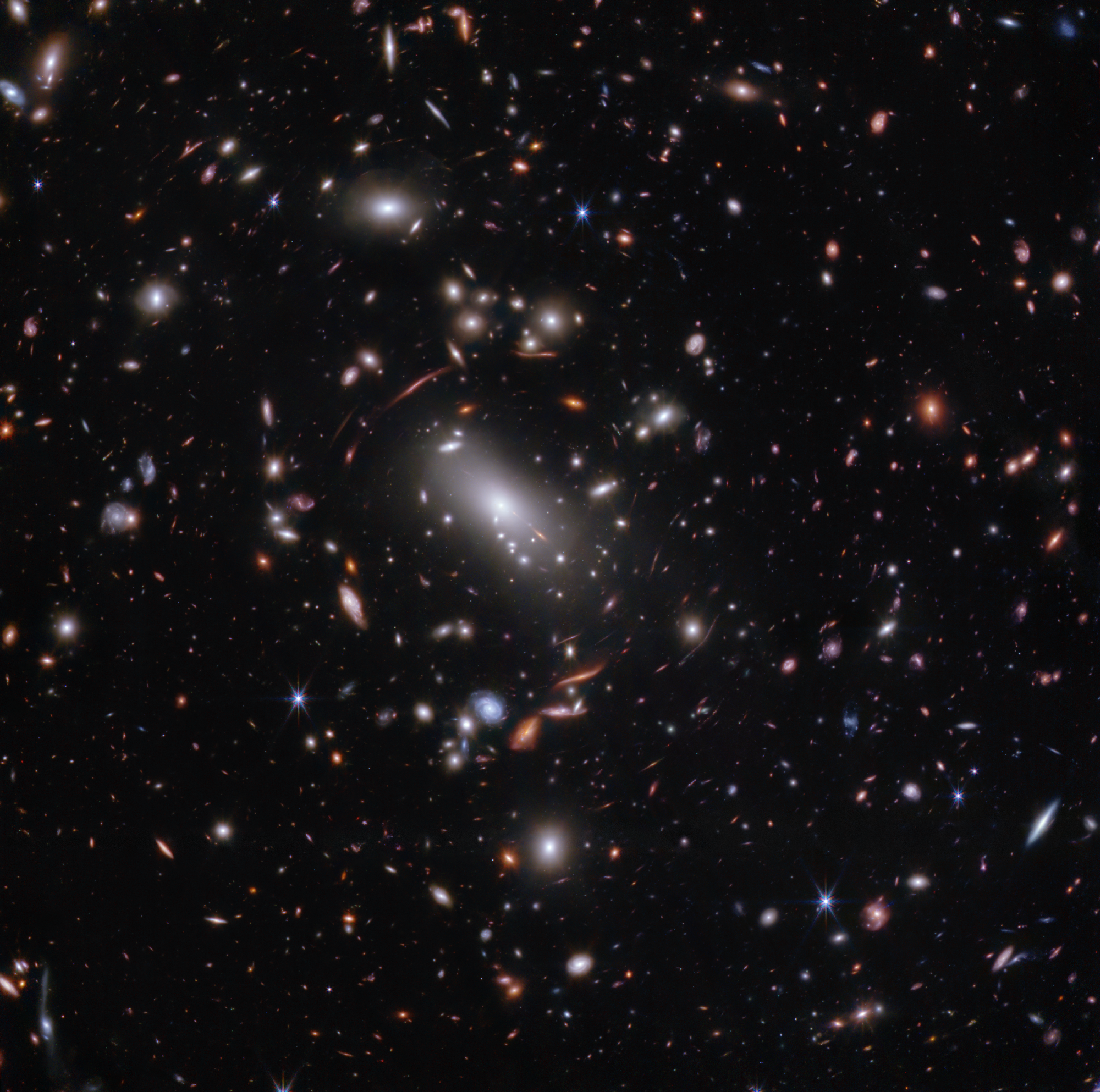 Thousands of overlapping objects at various distances are spread across this field, including galaxies in a massive galaxy cluster and distorted background galaxies behind the galaxy cluster. The background of space is black. The galaxies’ colors vary. The majority appear white or pink, some are shades of orange or blue. Most galaxies appear as fuzzy ovals, but a few have distinctive spiral arms. The most distant galaxies are the tiniest and appear as red dots or smudges. Several foreground stars with eight diffraction spikes appear as large as some of the smaller galaxies. Just above center is a very bright white, oversized oval, angled at 45 degrees and pointing to the top left and bottom right. This is a supergiant elliptical galaxy. Immediately around it are many thin, long, orange or pink arcs. They follow invisible concentric circles that curve around the center. These are background galaxies that have been stretched and distorted.