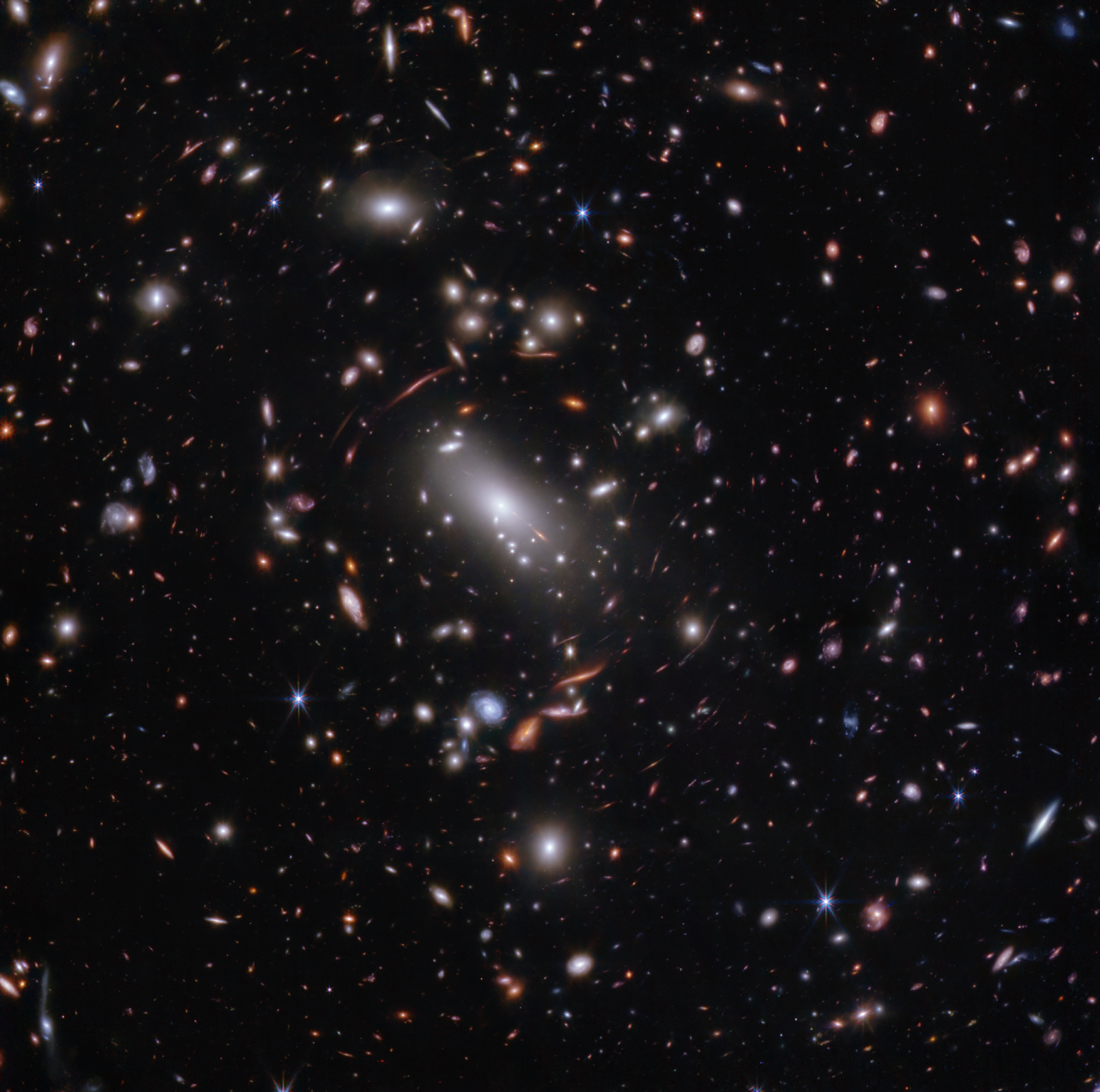 Thousands of overlapping objects at various distances are spread across this field, including galaxies in a massive galaxy cluster and distorted background galaxies behind the galaxy cluster. The background of space is black. The galaxies’ colors vary. The majority appear white or pink, some are shades of orange or blue. Most galaxies appear as fuzzy ovals, but a few have distinctive spiral arms. The most distant galaxies are the tiniest and appear as red dots or smudges. Several foreground stars with eight diffraction spikes appear as large as some of the smaller galaxies. Just above center is a very bright white, oversized oval, angled at 45 degrees and pointing to the top left and bottom right. This is a supergiant elliptical galaxy. Immediately around it are many thin, long, orange or pink arcs. They follow invisible concentric circles that curve around the center. These are background galaxies that have been stretched and distorted.