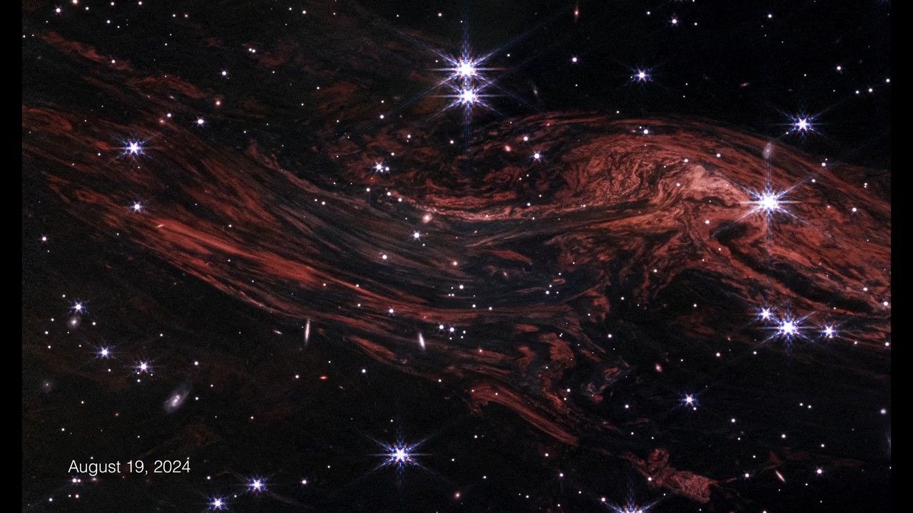 A few dozen white stars, some with Webb’s signature 8-point diffraction spikes, on the black background of space. Tightly packed, red-orange filaments extend horizontally from upper left to lower right, with a bulging upward curve on the right that is closer to the top. The filaments resemble wood grain. The image is labeled August 19, 2024 at bottom left.