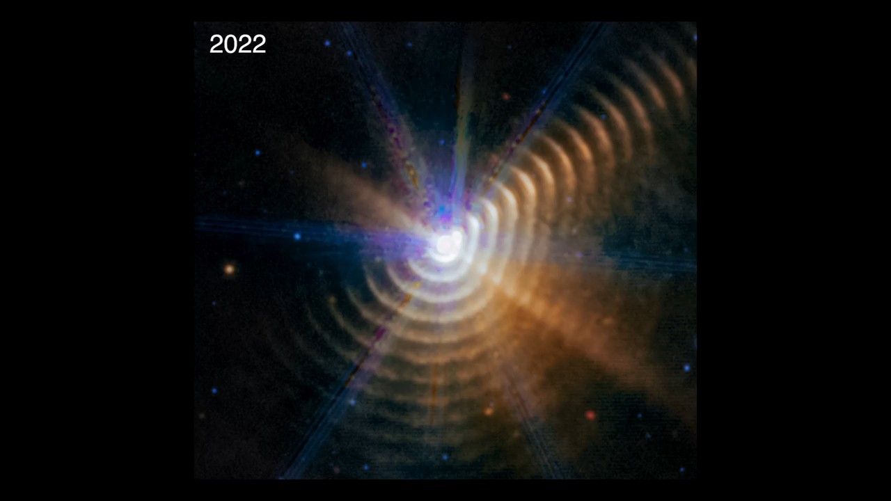 Wolf-Rayet 140 observed in 2022. A bright white point of light is surrounded by 17 regularly spaced, hazy dust shells at the bottom, right, and upper right, which look like tree rings. There is noticeably less color in the upper left. The central point, where the two stars are located, has a rough hexagon shape. There are several blue diffraction spikes. This area is toward the top right, where the outer rings appear bright orange.